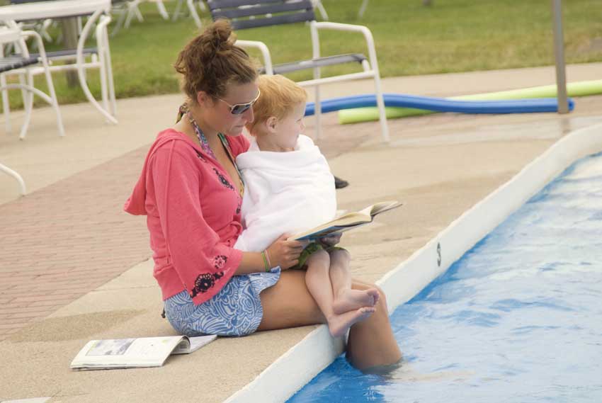 Woman reads to boy by the pool. Noodles in background. He's distracted.